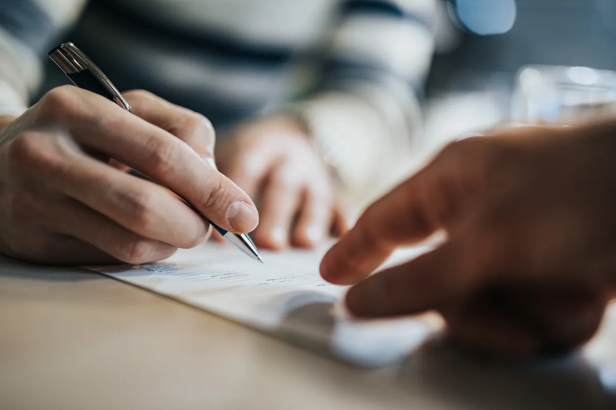 A hand pointing to a section of a document while someone else's hand signs the document with a pen.
