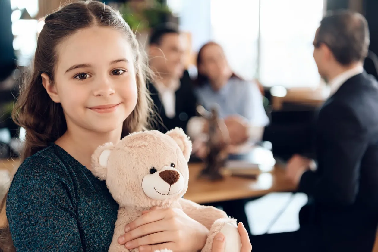 A young girl holding a teddybear and smiling wile two adults speak with a lawyer in the background.
