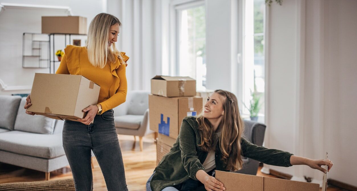 Two women in a large room with many moving boxes. The woman on the left is standing holding a box while the woman on the right squats and opens another box.