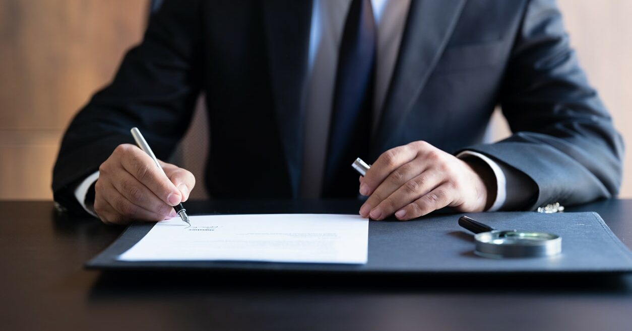 A person in a suit holding a pen over an official document. A magnifying glass lays next to the document.