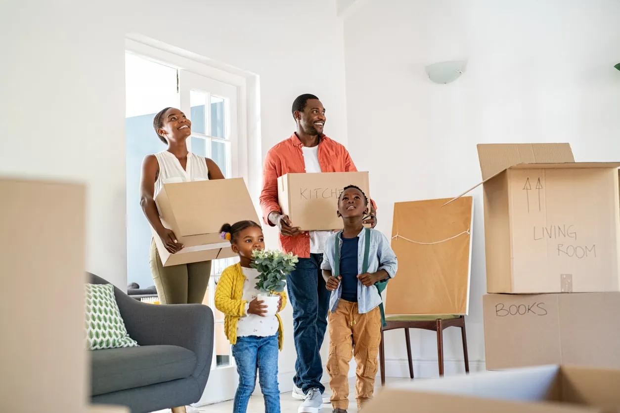 A family of four with a mother, father, son, and daughter walking into a room with white walls and several large cardboard boxes. The mother and father each have a cardboard box and the daughter is carrying a house plant.