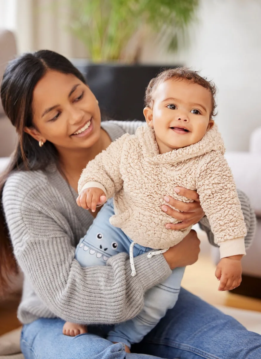 A smiling woman holding her daughter who is a toddler in a wool sweater.