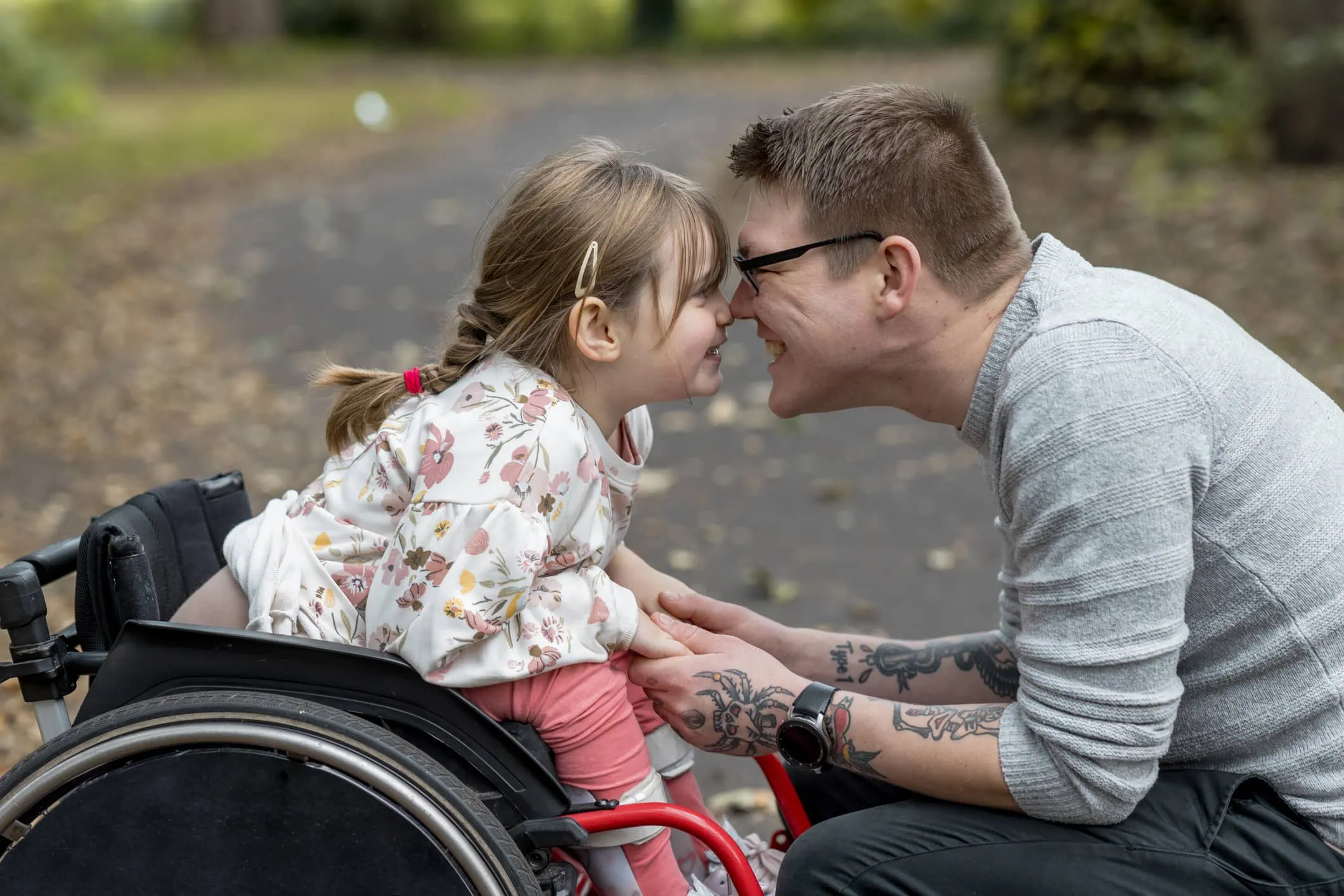 A father and his young daughter who is a wheelchair user pressing their noses together and smiling.