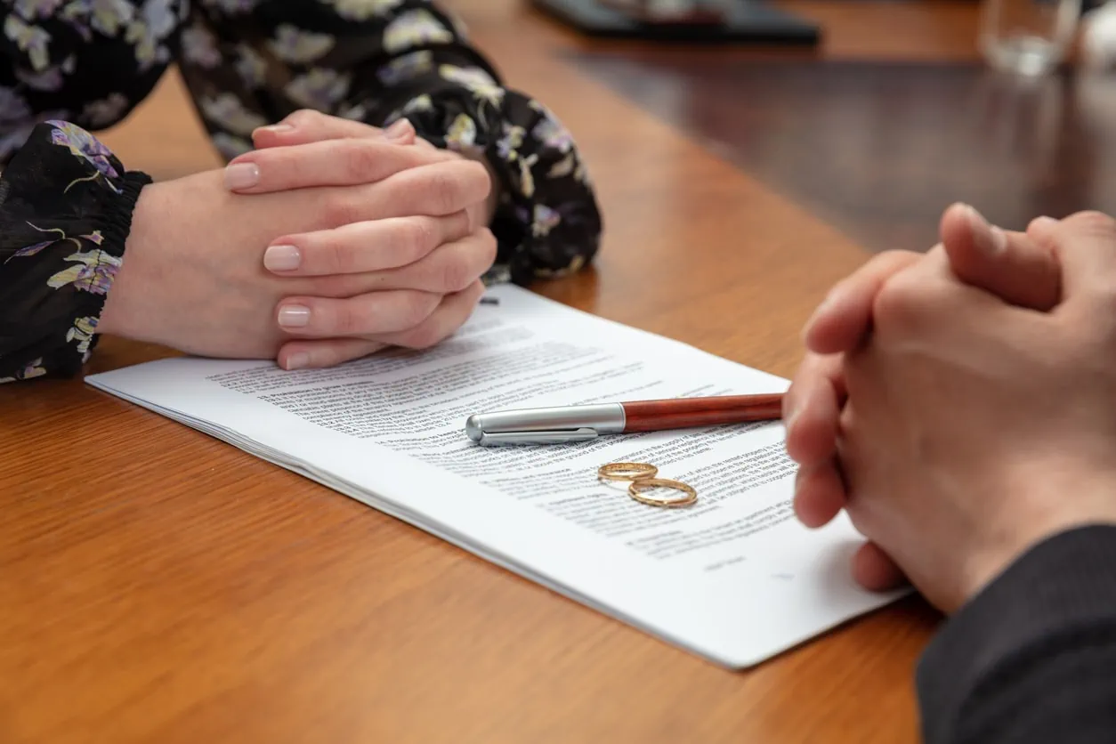 Two people with their hands folded on top of a legal document next to two wedding rings.