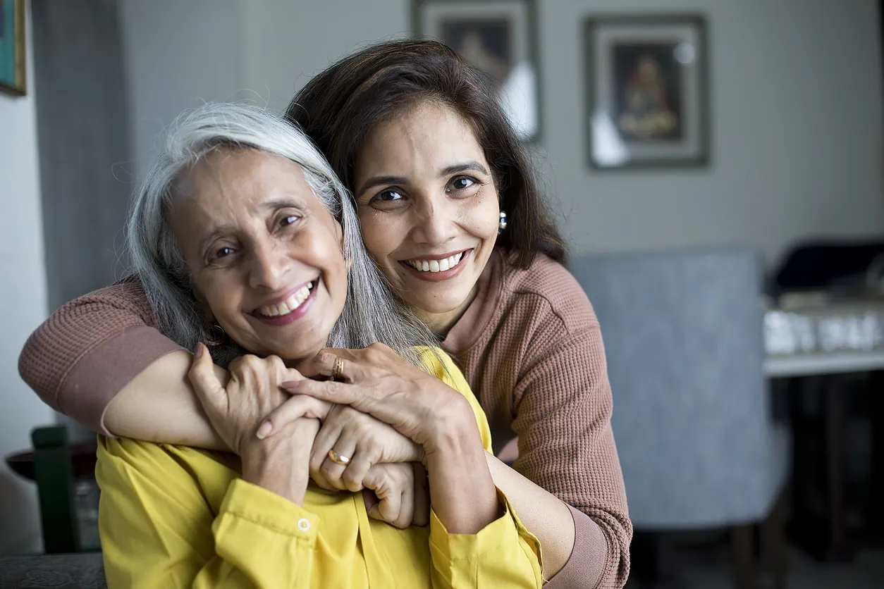 A middle-aged woman smiling and hugging a senior woman from behind.