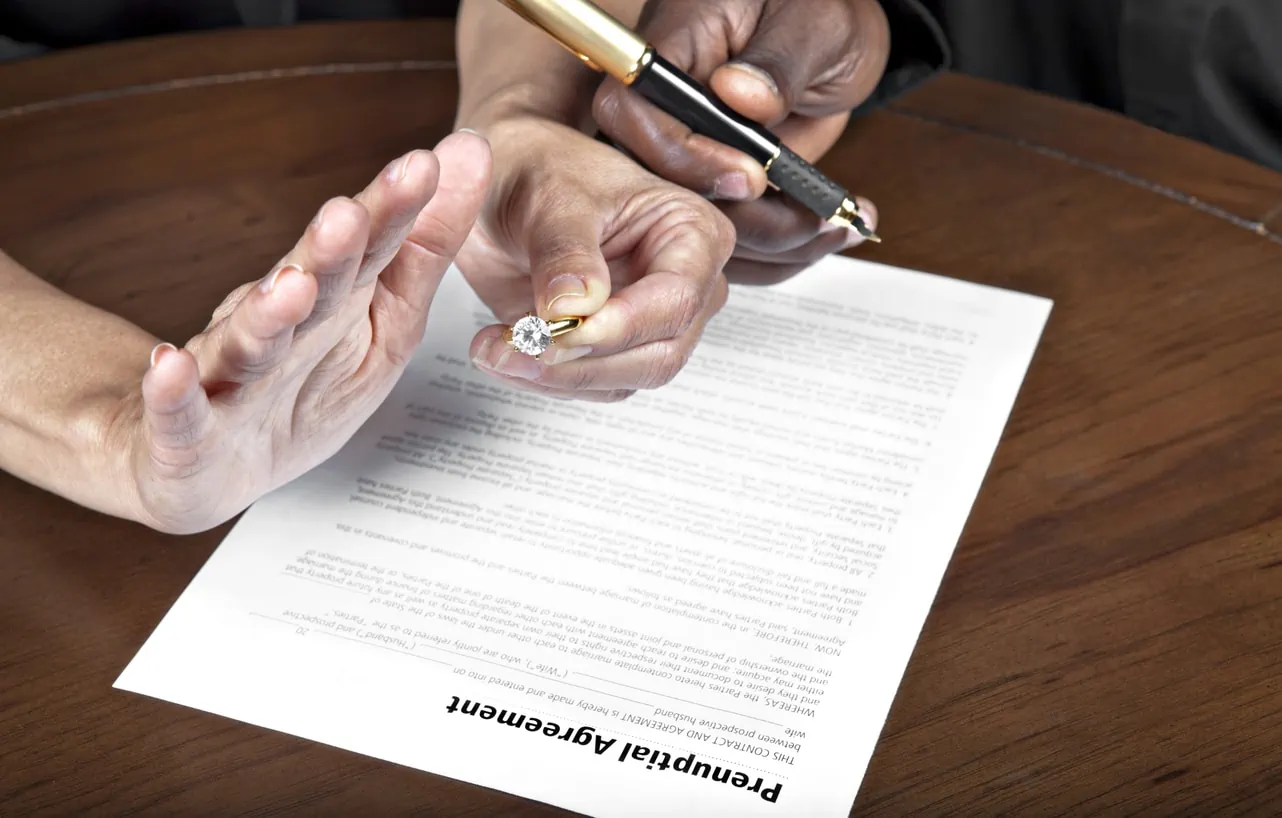 A person with one hand holding a diamond ring and another hand held out in a stopping gesture. Another hand attempts to hand them a pen. A prenuptial agreement sits on a table below.